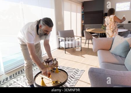 Mature man placing cake on coffee table while senior woman holding flute at home, copy space Stock Photo