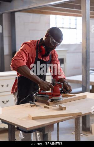 Red electric sander smoothing wooden plank on workbench in woodshop with offcuts Stock Photo