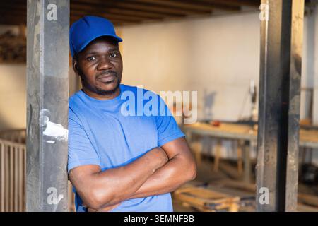 African American man leaning on metal beam crossing arms near workbench in workshop, copy space Stock Photo