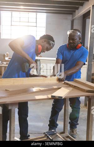 African American coworkers using power drill securing plank in workshop wearing goggles, earmuffs Stock Photo