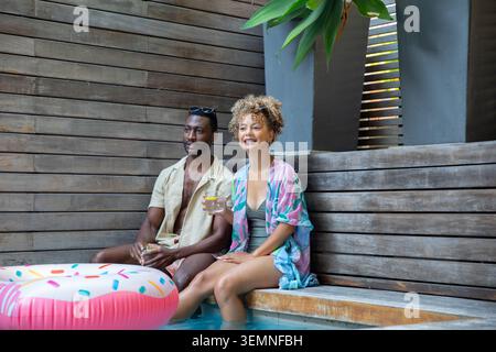 Diverse couple sitting on bench dipping feet in pool holding drinks near inflatable donut float Stock Photo