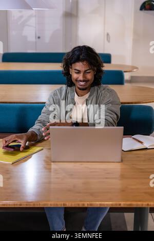 Indian man mid-20s smiling while working on silver laptop in shared study lounge with smartphone Stock Photo