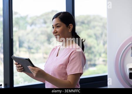 Adult female nurse in pink scrubs holding tablet, standing in clinic near window and pink device Stock Photo