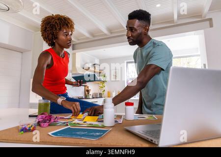 African American couple organizing printed papers and color swatches on kitchen island with laptop. Workspace, collaboration, documents, corkboard, ta Stock Photo