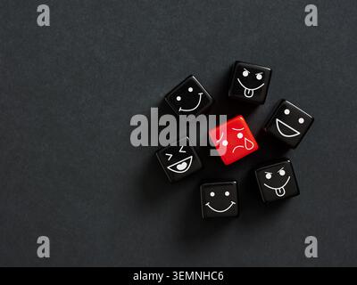 Red dice with a sad face surrounded by black dices showing various happy emotions on a dark background. Exclusion, bullying and loneliness concepts. Stock Photo