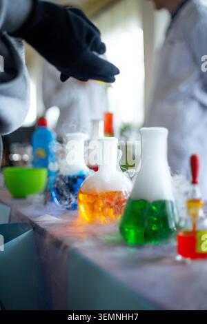 Students participate in a chemistry experiment with various colored liquids. They add substances to flasks while observing reactions. Smoke is visible Stock Photo