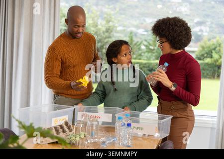 Diverse family sorting recycling with child at dining table near window bins PLASTIC GLASS PAPER Stock Photo