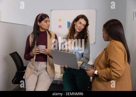 Three female staff sitting and standing around table holding laptop, tablet, pointing at whiteboard Stock Photo
