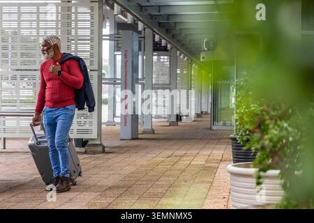 Senior African American man standing at CTICC concourse in red sweater holding suitcase, copy space Stock Photo