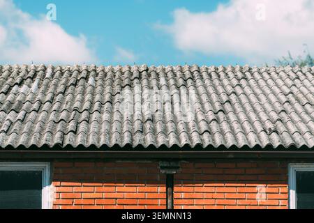A weathered corrugated tile roof crowns a red brick house under a bright blue sky with soft white clouds, highlighting architectural texture, pattern, Stock Photo