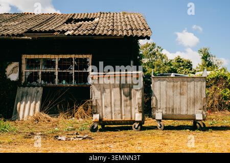Two weathered, rusty metal dumpsters on wheels standing on dry grass in front of an abandoned dark building with broken windows and damaged tiled roof Stock Photo