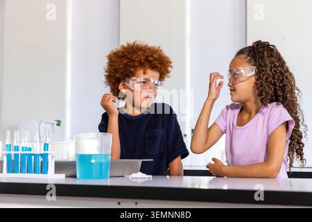 Two children wearing safety goggles mixing blue liquid in test-tube rack at school bench Stock Photo
