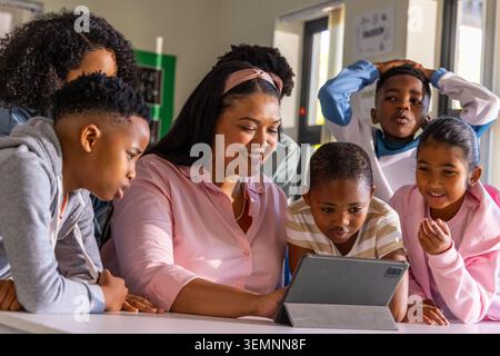 Female teacher wearing pink blouse and headband guiding schoolchildren using tablet in classroom Stock Photo
