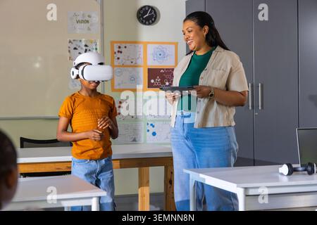 African American teacher guiding child wearing white VR headset at classroom table holding tablet Stock Photo