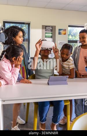 Diverse children gathering around classroom table seated child adjusting white VR headset, tablet Stock Photo