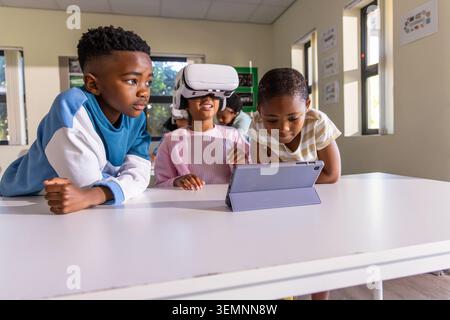 African American children trying virtual reality and using tablet at classroom table with headset Stock Photo