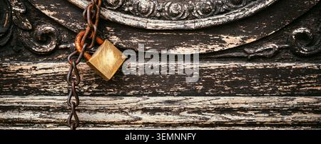 Close-up of a vintage brass padlock hanging on a rusty chain against an ornate, weathered wooden door with peeling paint and textured metalwork, evoki Stock Photo