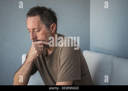 Pensive middle-aged man sitting on sofa deep in thought looking worried and contemplative, selective focus Stock Photo