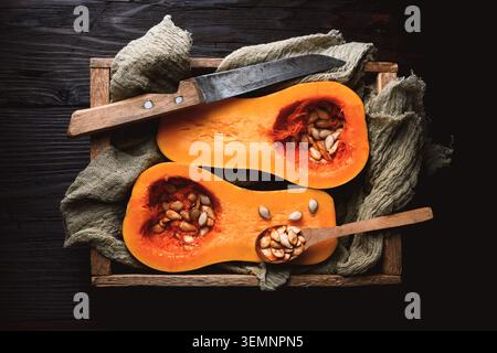 Top-down view of butternut squash halves with pumpkin seeds, spoon, and knife on wood surface. Rustic food scene for autumn recipe or harvest ingredient styling Stock Photo
