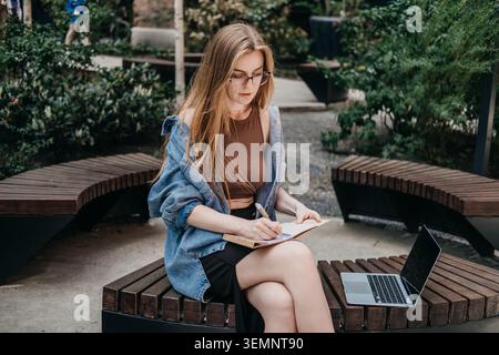 Woman writes in notepad near laptop while sitting on wooden public bench outdoors. Digital nomad life, laptop on bench, modern work solutions, Tech Stock Photo