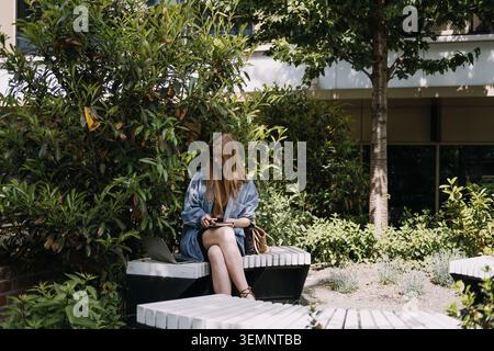 Woman sits on bench in green courtyard writing in notebook next to laptop computer. Connected work lifestyle, mobile technology productivity, healthy Stock Photo