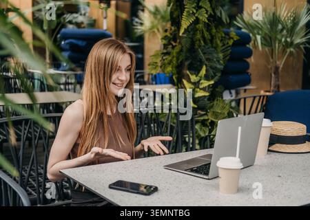 Young woman gestures and smiles while using laptop at outdoor cafe table. Female mentor teaching online, virtual education instructor, educational Stock Photo