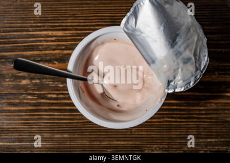 Open plastic cup of strawberry yogurt with a spoon on wooden background. Top view of pink fruit yogurt in an opened container with a metal spoon and f Stock Photo
