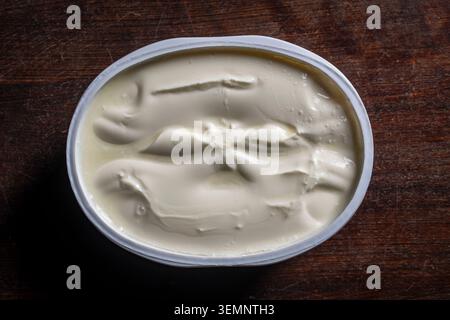 Creamy white sour cream or processed cheese in plastic container on wooden table, close up. Top view of thick white yogurt cream cheese or sour cream Stock Photo