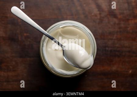 Fresh organic yogurt in a glass jar with a spoon on wooden background. Top view of creamy white yogurt or sour cream inside a glass jar with a metal s Stock Photo