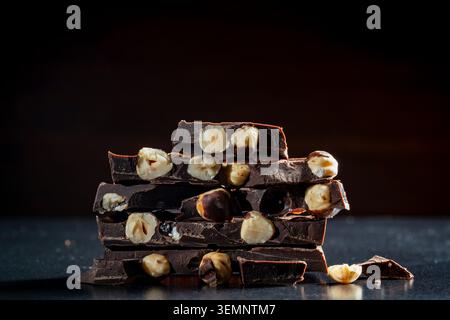 Stack of broken dark chocolate bars with whole hazelnuts. Close up of dark chocolate pieces with whole hazelnuts stacked on a black background Stock Photo