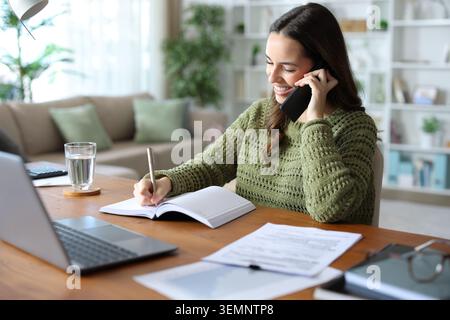 Happy teleworker making phone call and writing in agenda on a wooden table Stock Photo