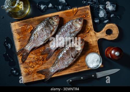 Fresh raw fish crucian carp with ingredients on wooden cutting board, close up. Top view of three raw silver fish on a wooden board with ice cubes, ol Stock Photo