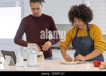 African American couple baking pastry at counter, pressing dough into pie dish, tablet and apron Stock Photo
