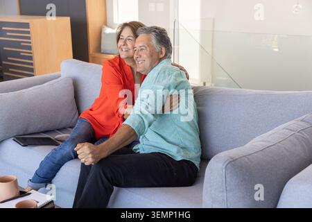 Senior woman and mature man sitting on light gray sofa hugging, smiling, looking left with tablet Stock Photo