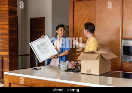 Two Asian men unpacking, arranging items on kitchen island with framed artwork, box, bubble wrap Stock Photo