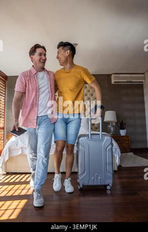 Diverse male partners standing, smiling and holding passports in hotel room with silver carry-on Stock Photo