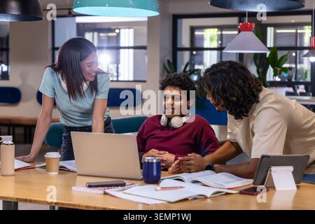 Diverse peers leaning over laptop, discussing notes at modern study lounge with headphones. Collaboration, teamwork, campus, coworking, notebooks, tex Stock Photo