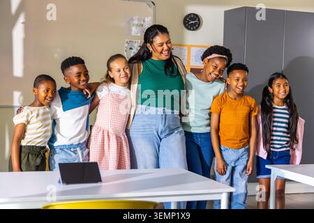 Diverse group of six children with female teacher smiling in classroom, tablet on table Stock Photo