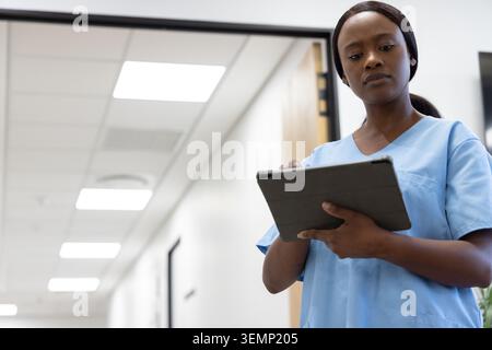 Woman holding tablet standing in hospital corridor wearing light blue scrubs, copy space Stock Photo