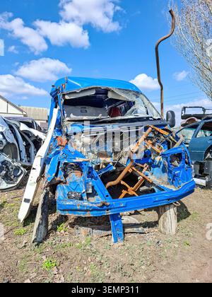 A dismantled turquoise car frame sits in an auto salvage yard. The engine and front bumper are missing, and the car is stripped for spare parts. Other Stock Photo