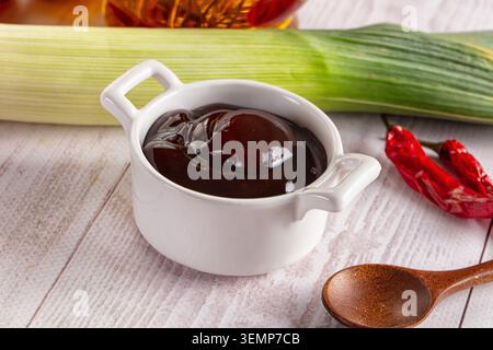 Glossy dark brown sauce in white ceramic bowl with handles, fresh green leek and dried red chili peppers on rustic wooden surface, wooden spoon, food Stock Photo