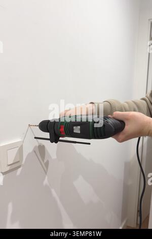 Hand using corded electric drill horizontally near light switch on white hallway wall Stock Photo