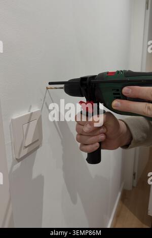 Person drilling pilot hole in white wall near light switch with green corded electric drill Stock Photo