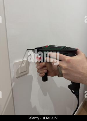 Male hand using corded electric drill horizontally to bore hole in white wall near light switch Stock Photo