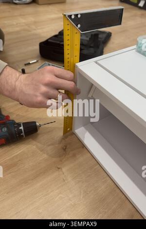 Carpenter using yellow square ruler measuring height of white shoe cabinet during furniture assembly Stock Photo