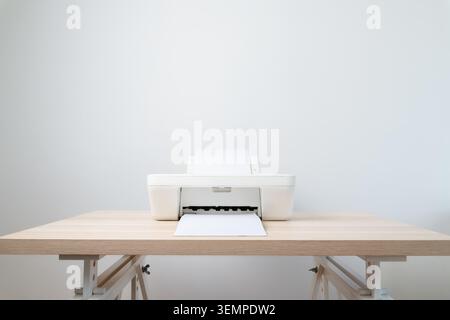 Minimal white printer sits on a light wooden desk with a sheet of paper feeding through, suggesting an organized modern office or home workspace focus Stock Photo