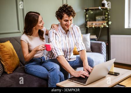 A happy young couple sits together on a comfortable sofa, browsing on a laptop in their living room. The woman enjoys a warm drink. Stock Photo