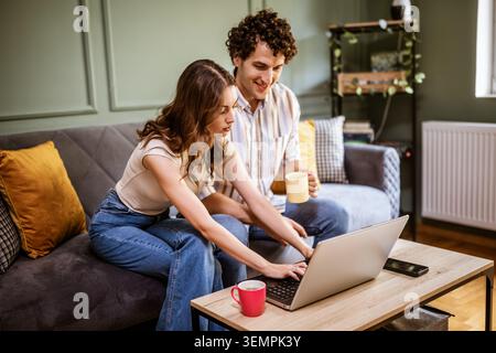 A smiling couple spends quality time at home, exploring content on a laptop. Their relaxed expressions and casual setting suggest online shopping or f Stock Photo