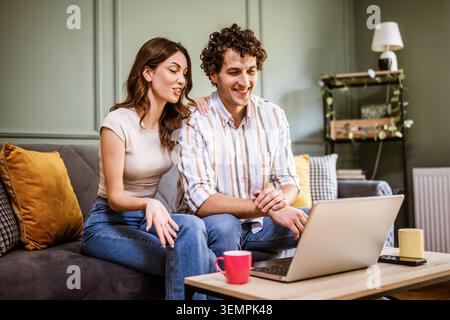 Adult couple is having video call with family. Man and woman sitting at sofa at home and using laptop for online communication. Stock Photo