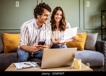 A woman holds a bill or statement while speaking with her partner on the sofa. Their are working together to understand expenses, manage finances, or Stock Photo